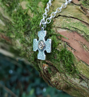 Side view of the Crux Quadrata, a Greek cross pendant with a gold spiral at the centre, hanging against mossy tree bark. Handmade in Ireland.