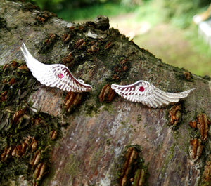 Handmade sterling silver Angel Wings Ear Cuff earrings with red gemstone set in the middle. The jewelry pieces are displayed on tree bark. These angel earrings are part of Elena Brennan's Irish My Angel jewelry collection