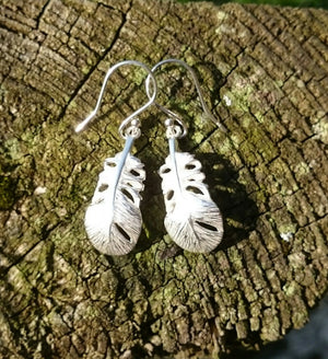The Baby Angel Feather Drop Earrings, made of sterling silver, are part of the My Angel Jewellery collection. The angel earrings are laid against tree bark.