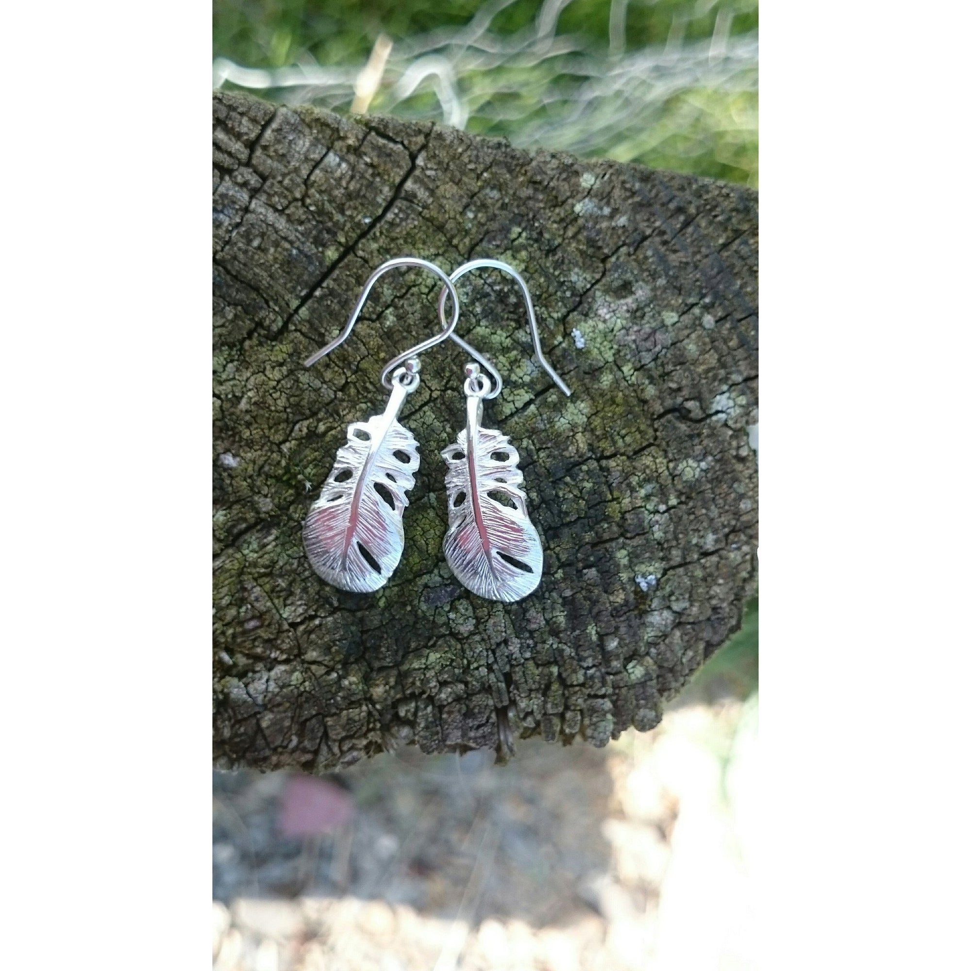 These Irish Baby Angel Feather Drop Earrings are laid against tree bark. They are part of Elena Brennan's My Angel jewellery collection.