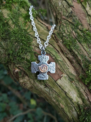 The Crux Quadrata, a Greek cross pendant with a Celtic gold spiral at the centre, hanging against mossy tree bark. Handmade in Ireland.