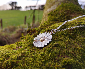 This Dog Daisy pendant is made of sterling silver and gold and is sitting on a mossy tree branch. Daisy Jewellery handmade in Ireland by Elena Brennan.